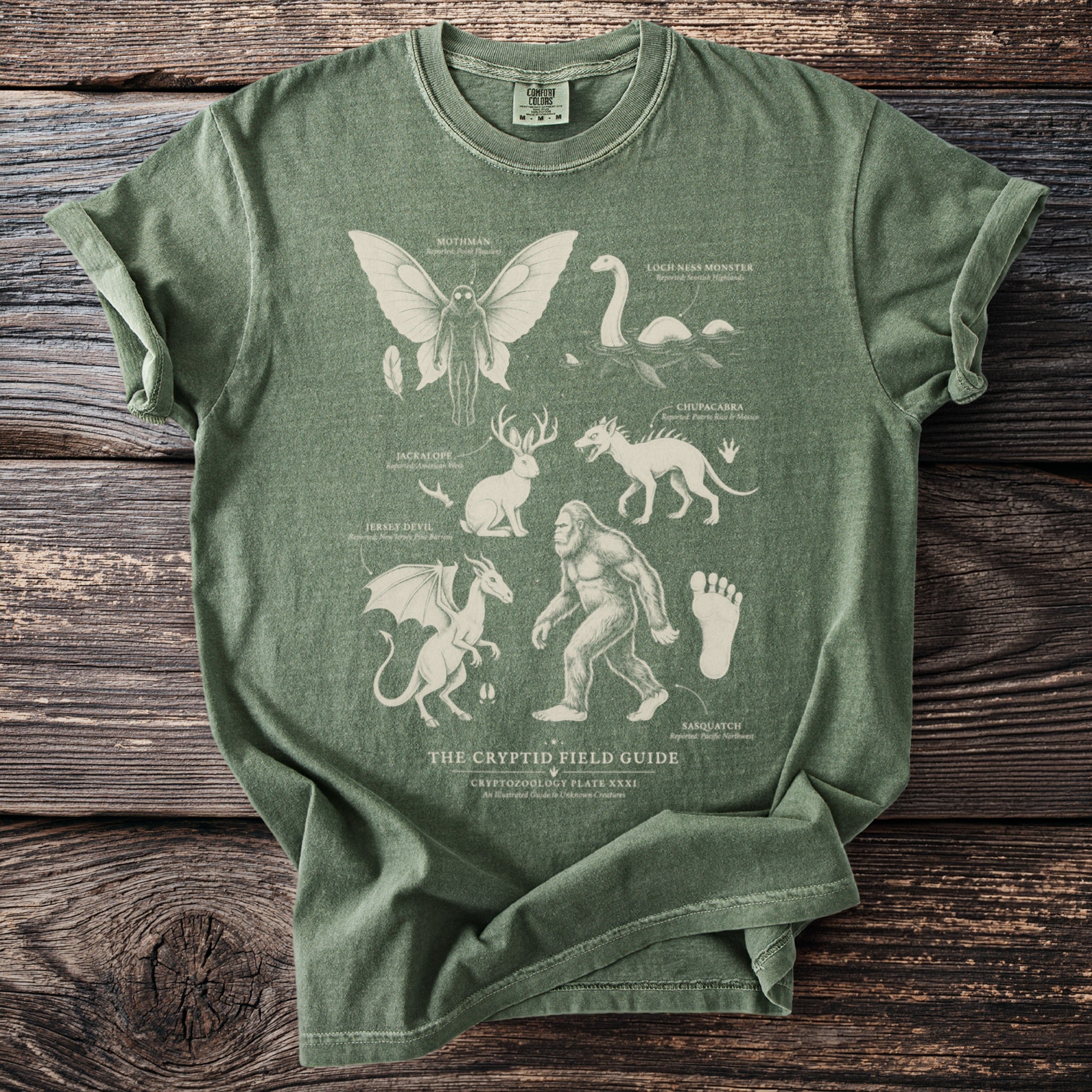 Green t-shirt featuring "The Cryptid Field Guide" with illustrations of Mothman, Loch Ness Monster, and Sasquatch.
