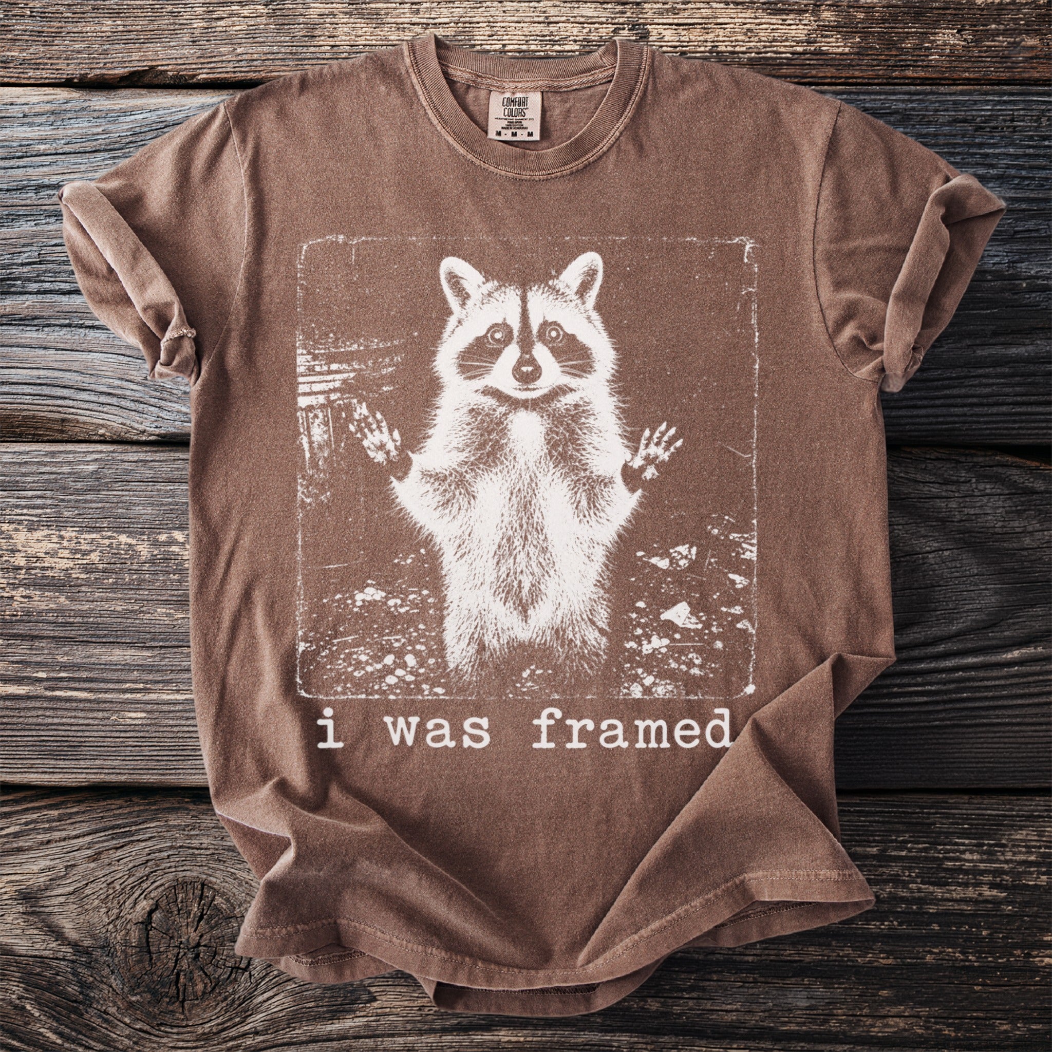 Brown t-shirt with white graphic of raccoon and text saying "i was framed."
