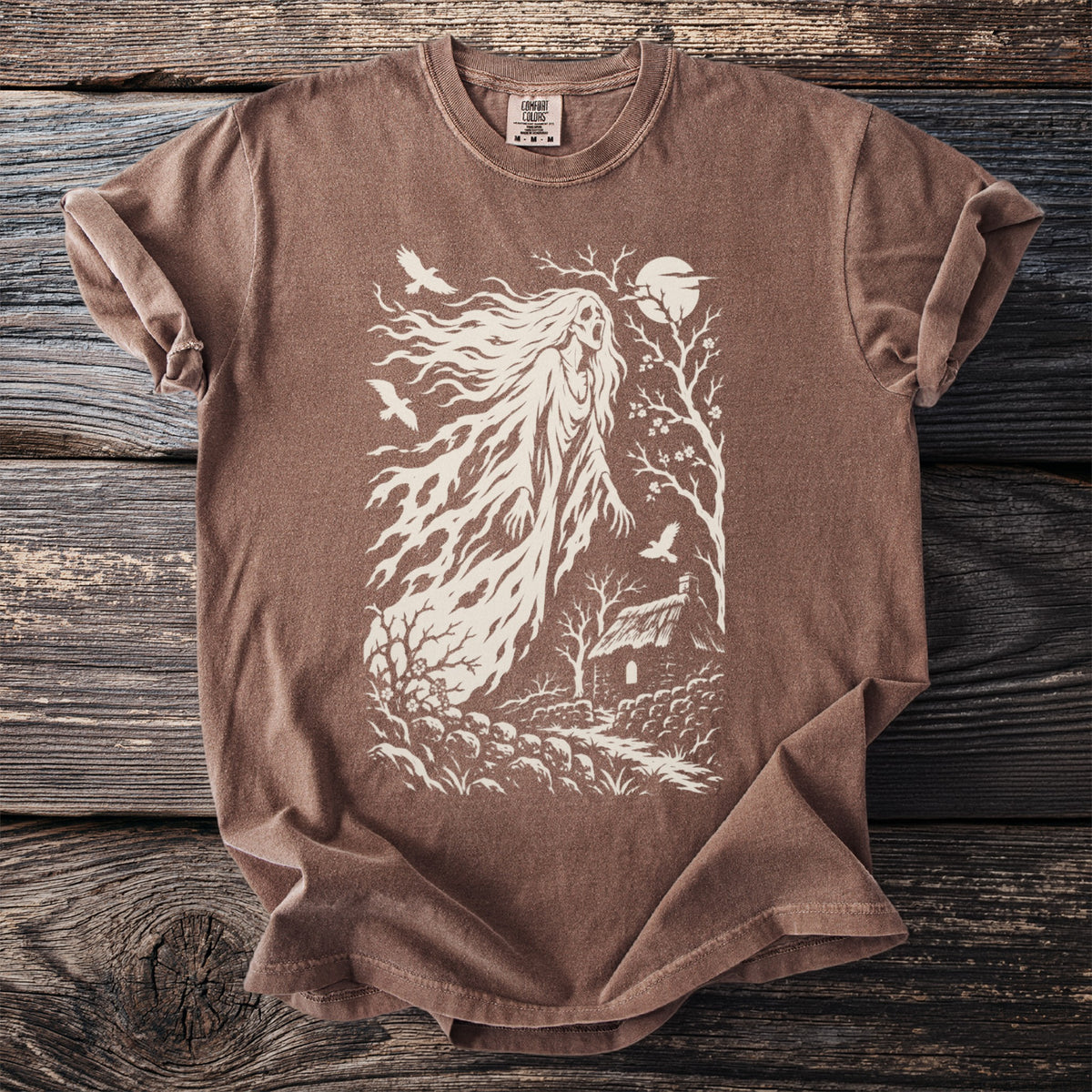 Brown t-shirt with a white graphic of a ghostly figure, a cottage, and birds.