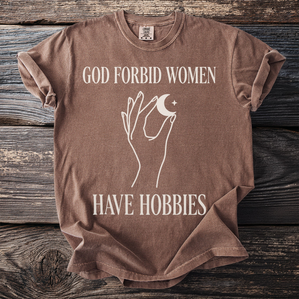 Brown t-shirt with white text "GOD FORBID WOMEN HAVE HOBBIES" and celestial graphic.