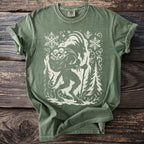 Green t-shirt with a cream Krampus silhouette carrying a sack and a staff among fir trees.