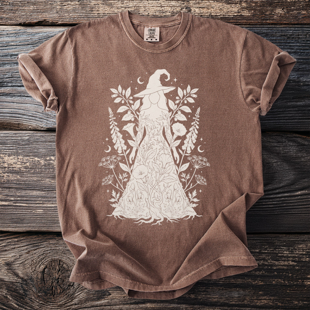 Brown t-shirt with a white line drawing of a witch figure wearing a floral dress.