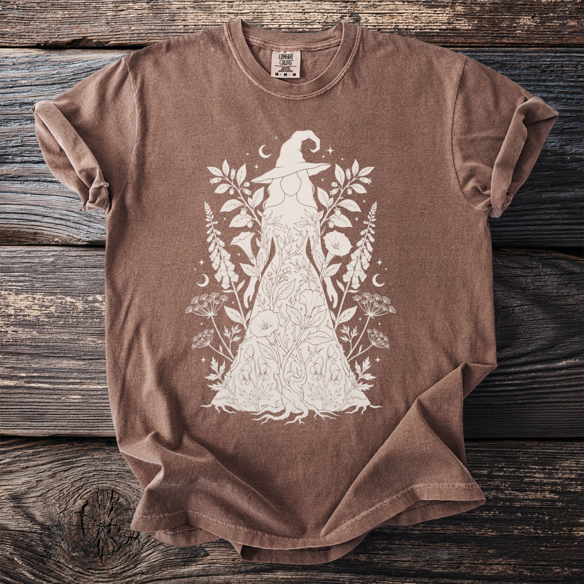 Brown t-shirt with a white line drawing of a witch figure wearing a floral dress.