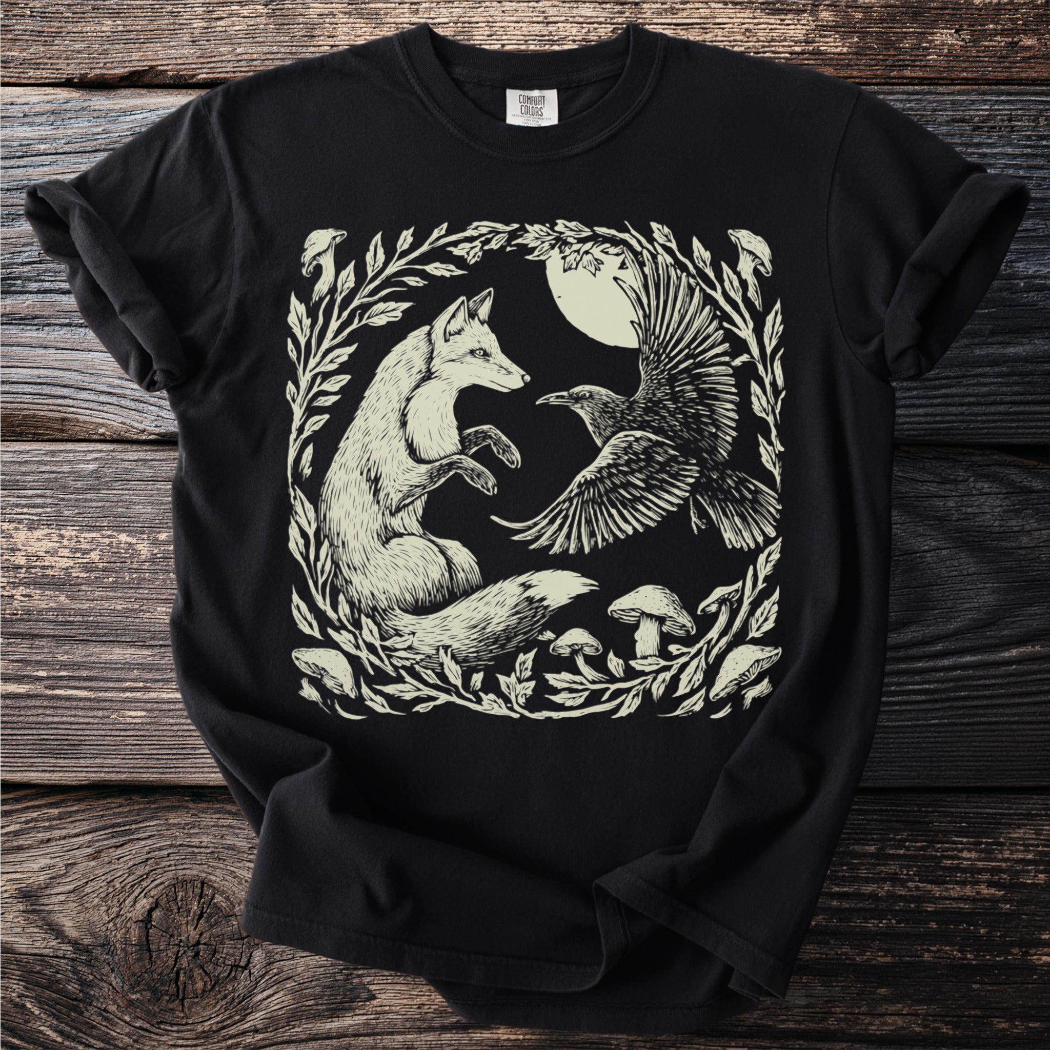 Black t-shirt with a cream linocut art of a fox and raven with moon and mushrooms.