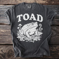 Dark grey t-shirt with distressed white print of "TOAD" above a toad, mushrooms, and insects.