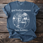 "God forbid women have hobbies" text on a blue t-shirt with library scene illustration.