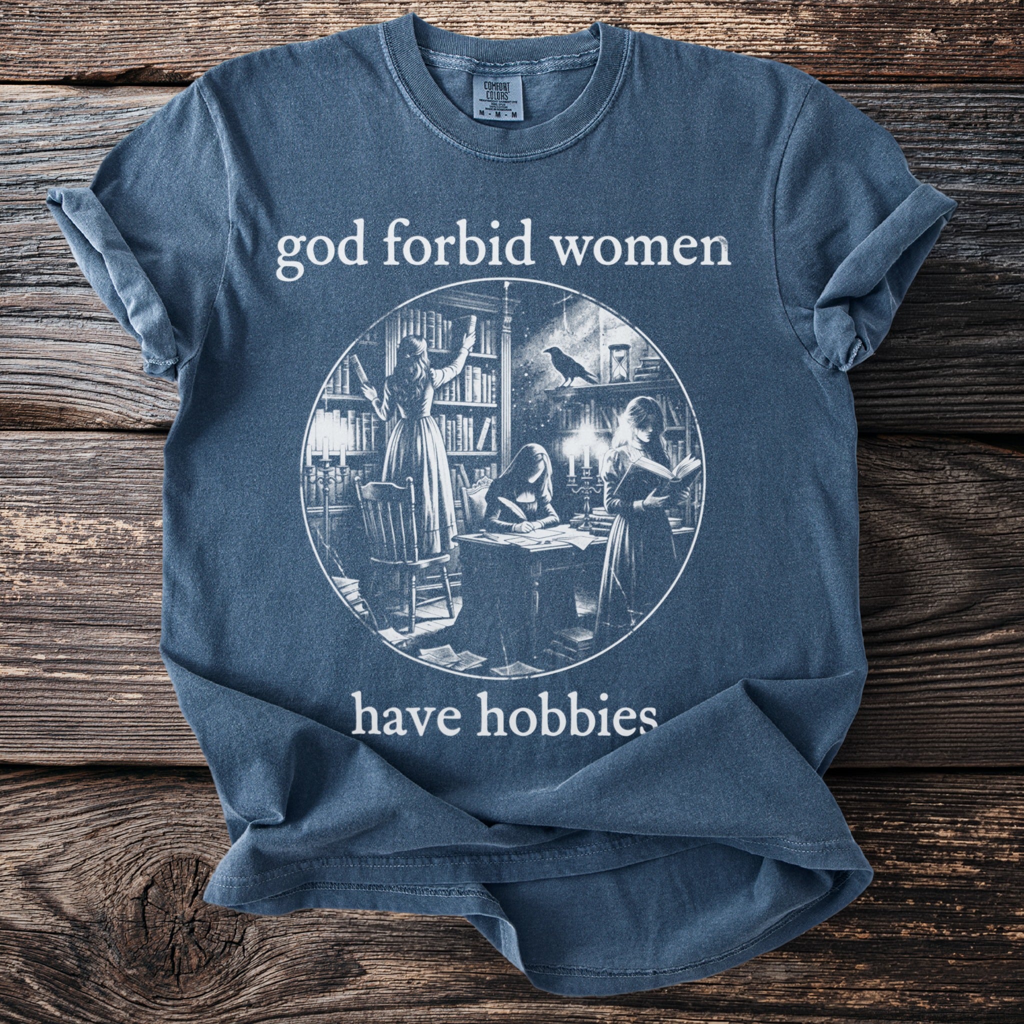 "God forbid women have hobbies" text on a blue t-shirt with library scene illustration.