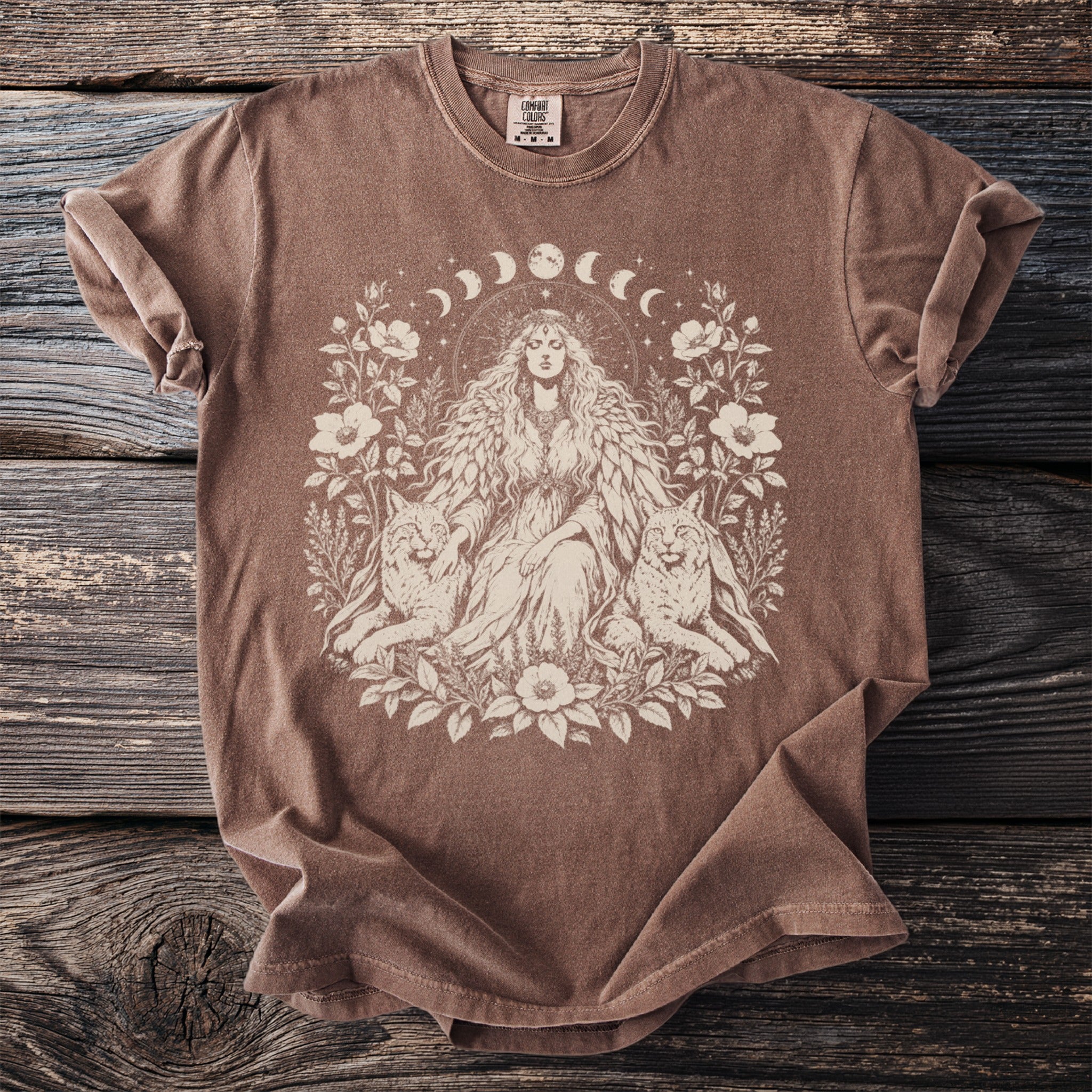 Brown Comfort Colors t-shirt with a cream graphic of a woman, two lynxes, and floral elements.