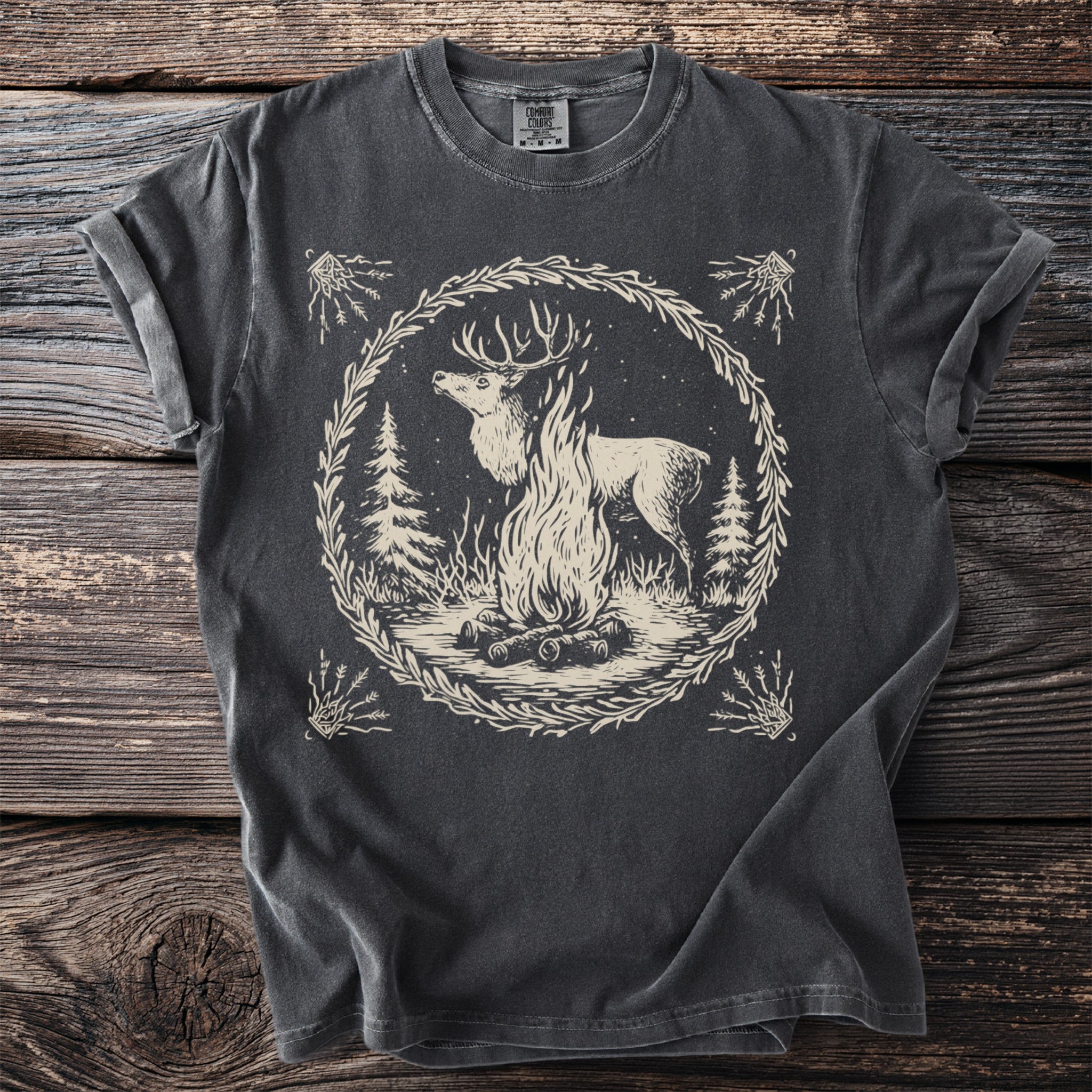 Dark t-shirt with a beige graphic of a deer by a campfire within a wreath.