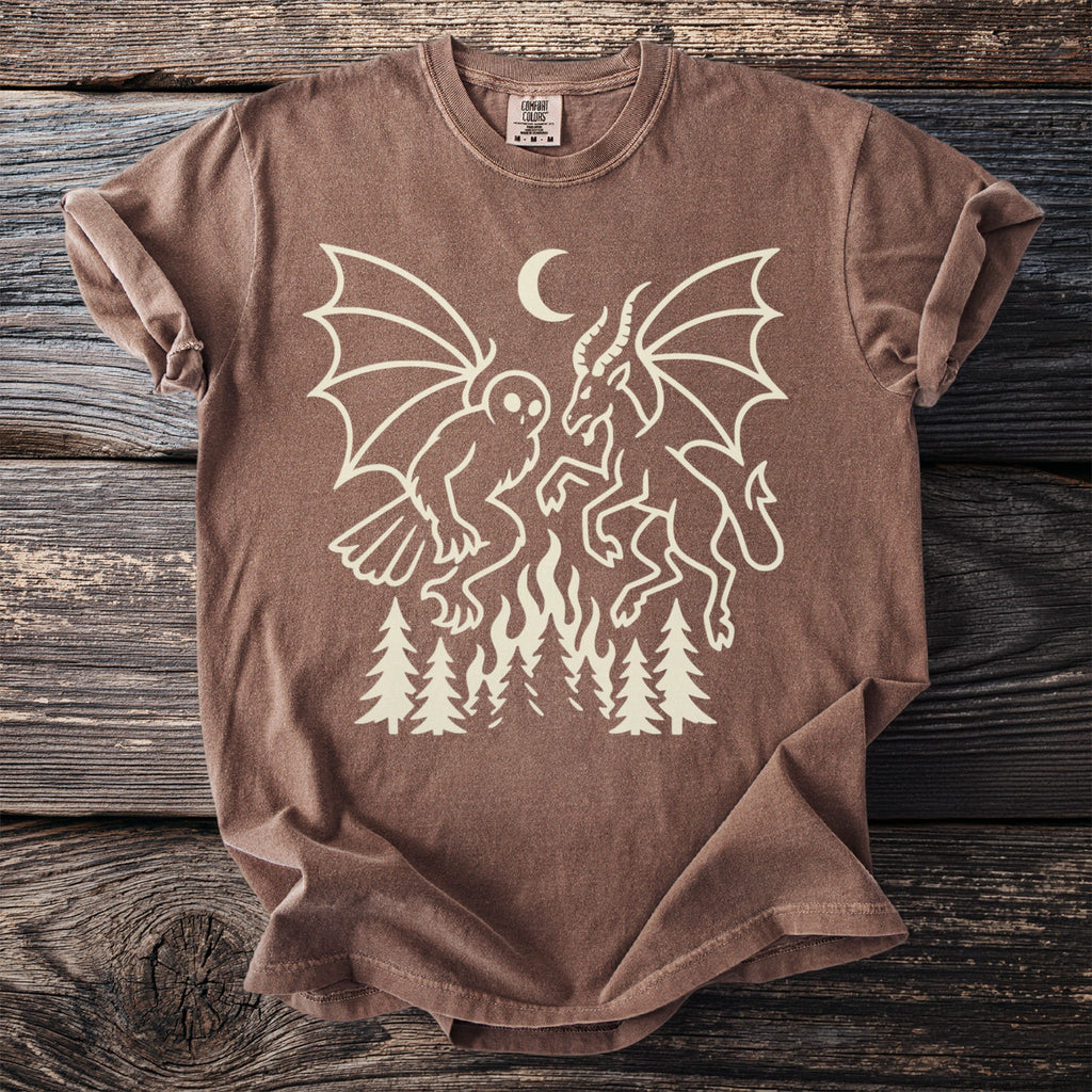 Brown t-shirt with cream linework of a creature and a goat-like figure over a fire and trees.