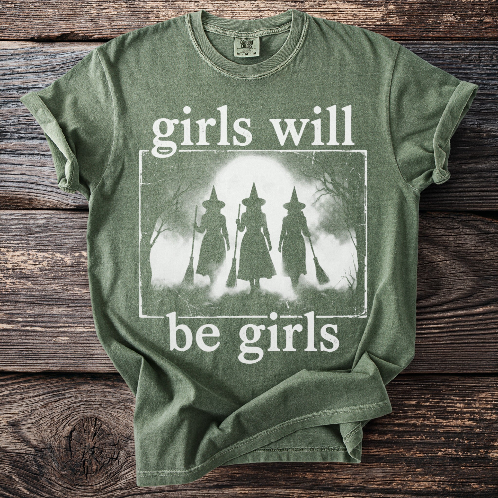 Green t-shirt with white distressed graphic of three witches and text "girls will be girls".