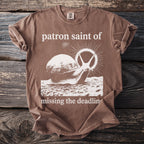 Brown t-shirt with a graphic of a snail and text reading "patron saint of missing the deadline."