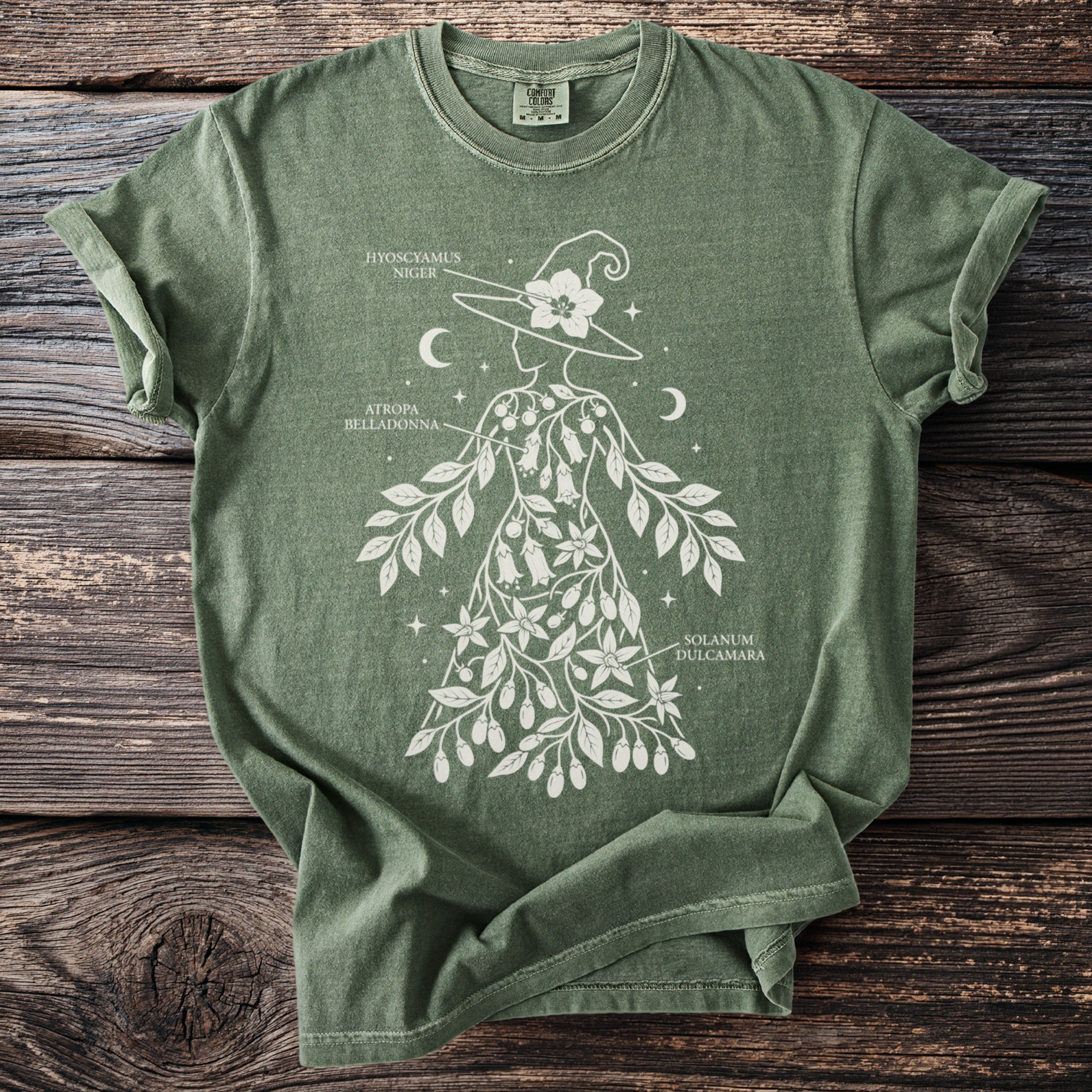 Green t-shirt with white botanical illustration of a witch and labeled plants.