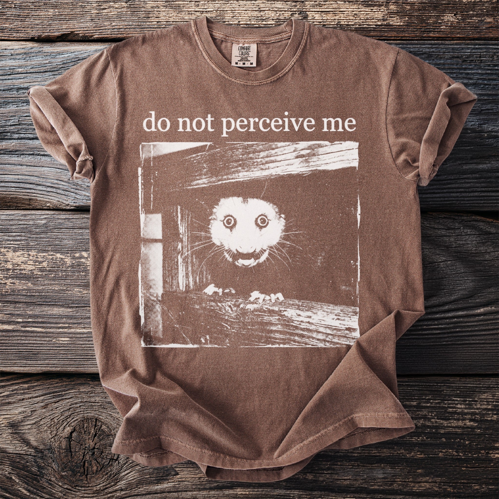 Brown T shirt with a bushbaby peering through wooden planks and “do not perceive me” text.
