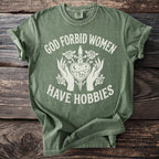 Green t-shirt with white text "God forbid women have hobbies" and graphic.