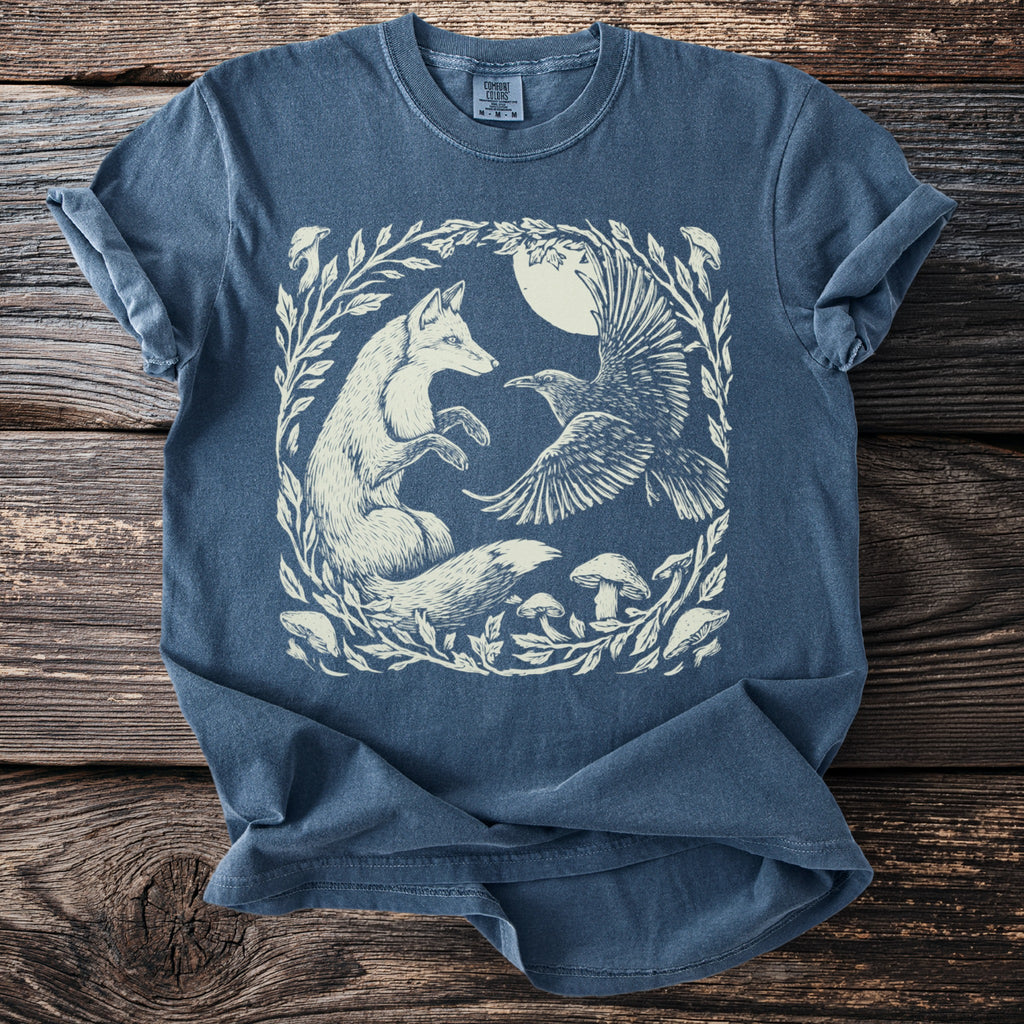 Blue t-shirt with a white illustration of a fox, raven, and mushrooms in a circular leafy frame.