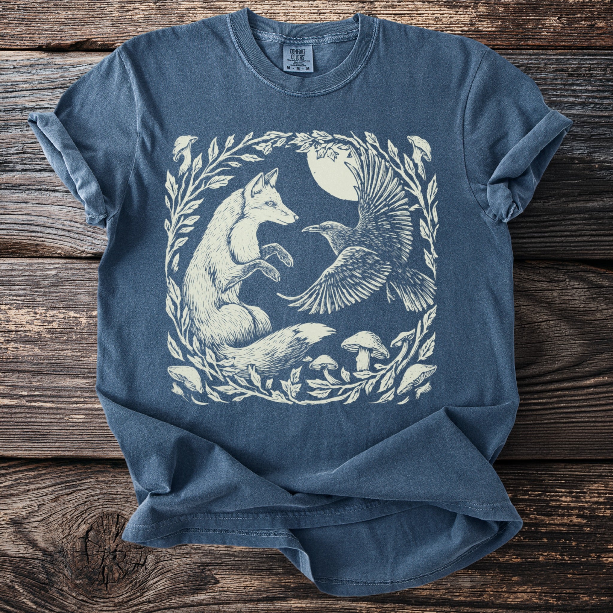 Blue t-shirt with a white illustration of a fox, raven, and mushrooms in a circular leafy frame.