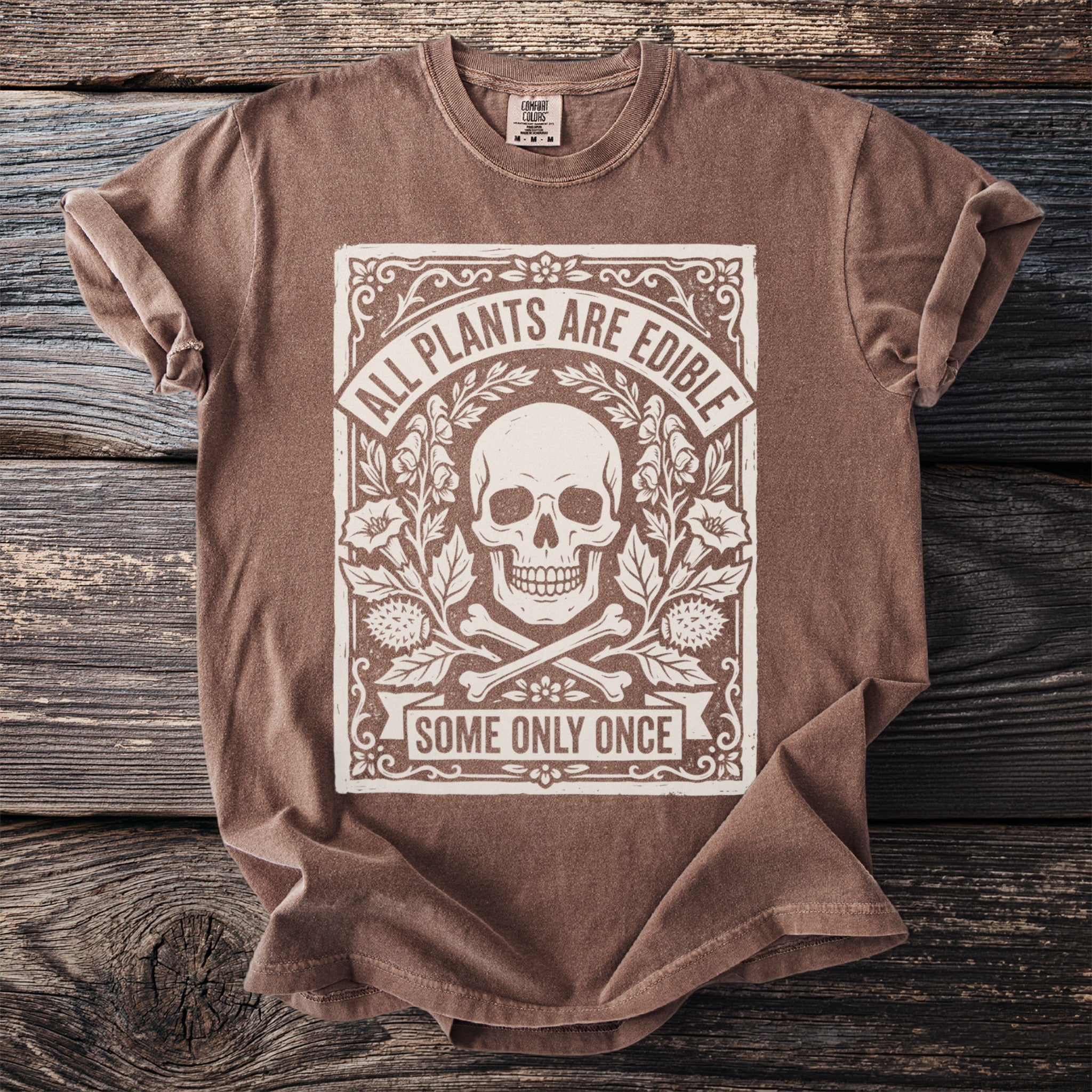 Brown t-shirt with skull, bones, plants, and text "ALL PLANTS ARE EDIBLE SOME ONLY ONCE".