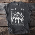 Dark grey t-shirt with white text "sunken city" and a kraken above three figures.
