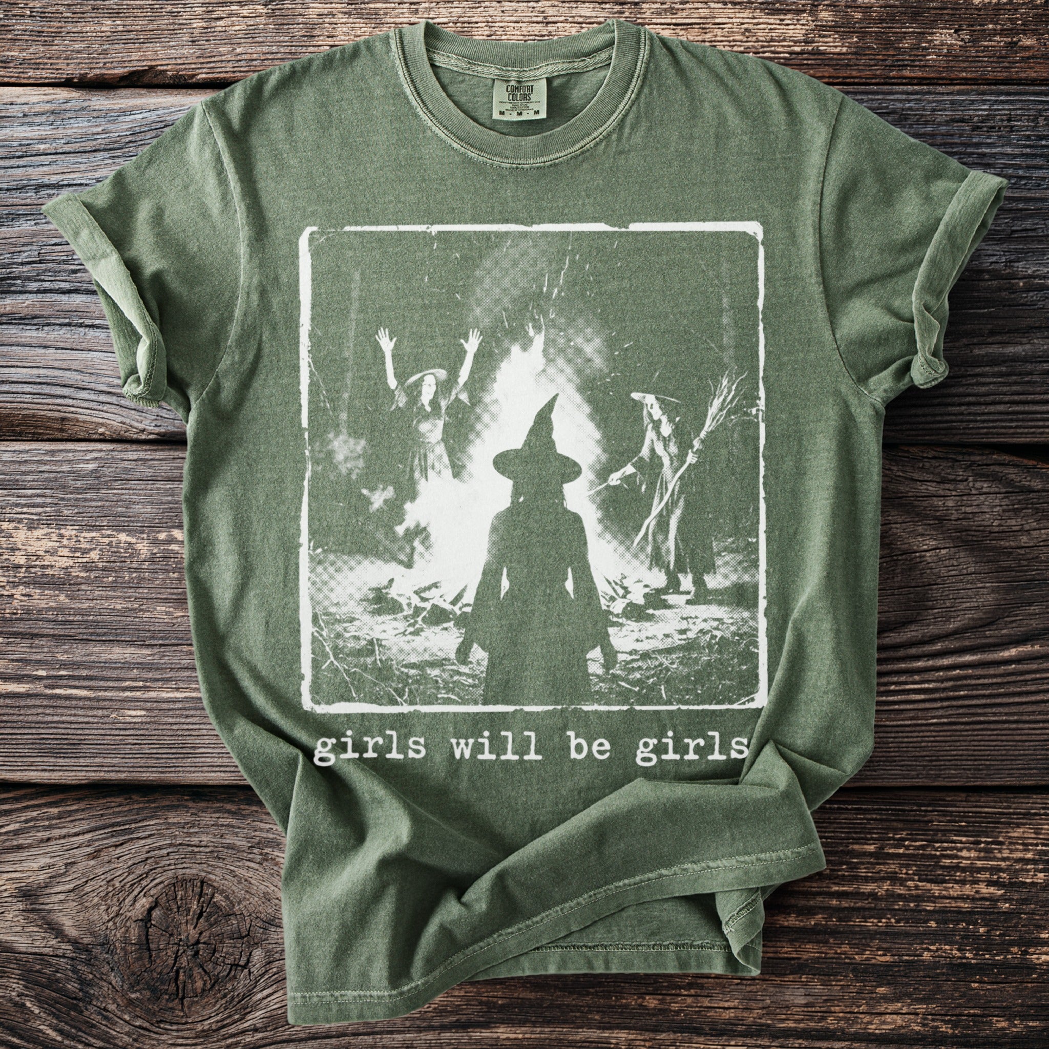 Green t-shirt with graphic of witches around a bonfire and text "girls will be girls".