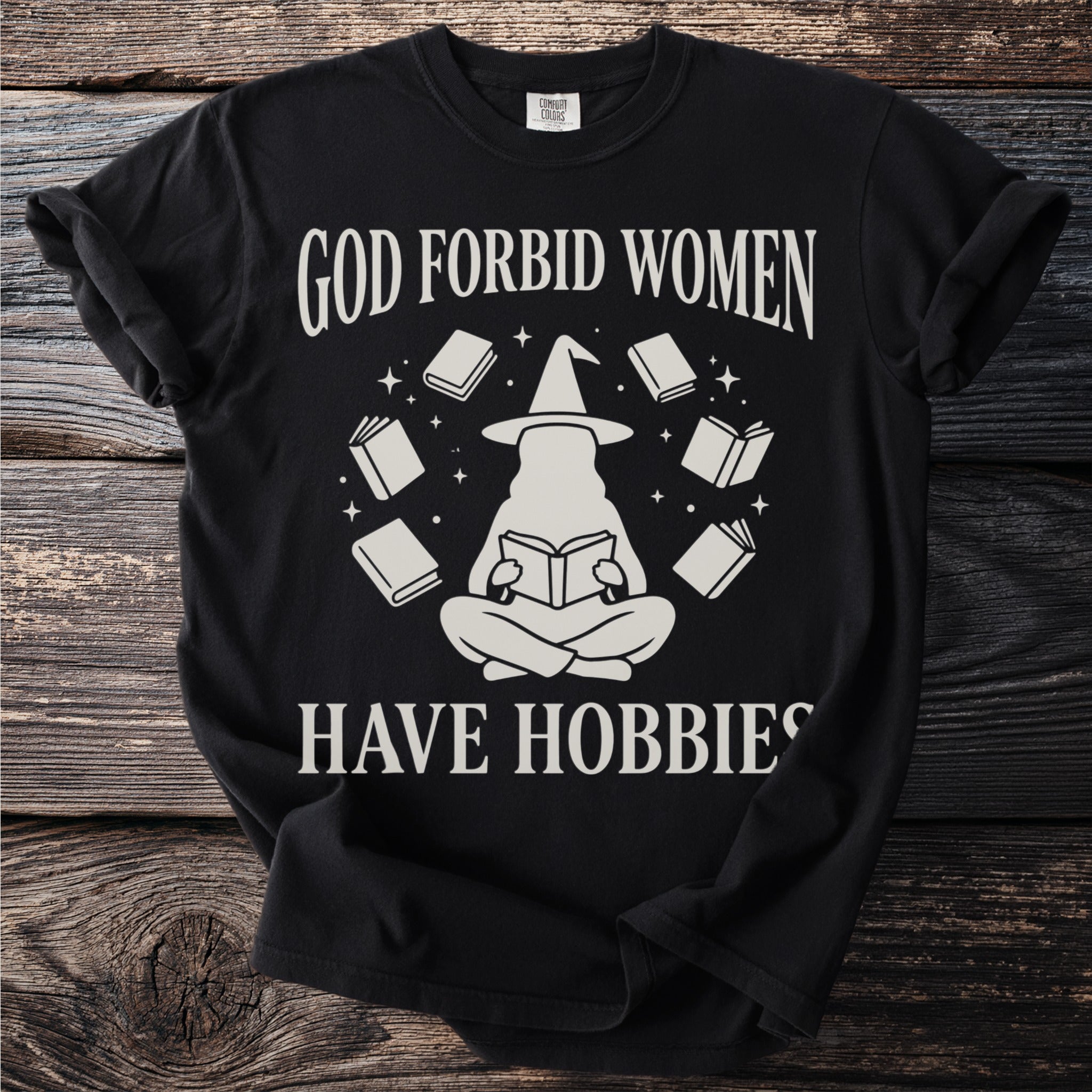 Black t-shirt with graphic reading "God forbid women have hobbies" and a witch reading a book.
