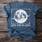 Blue t-shirt with graphic of three witches flying across a moon, text reads "girls will be girls".
