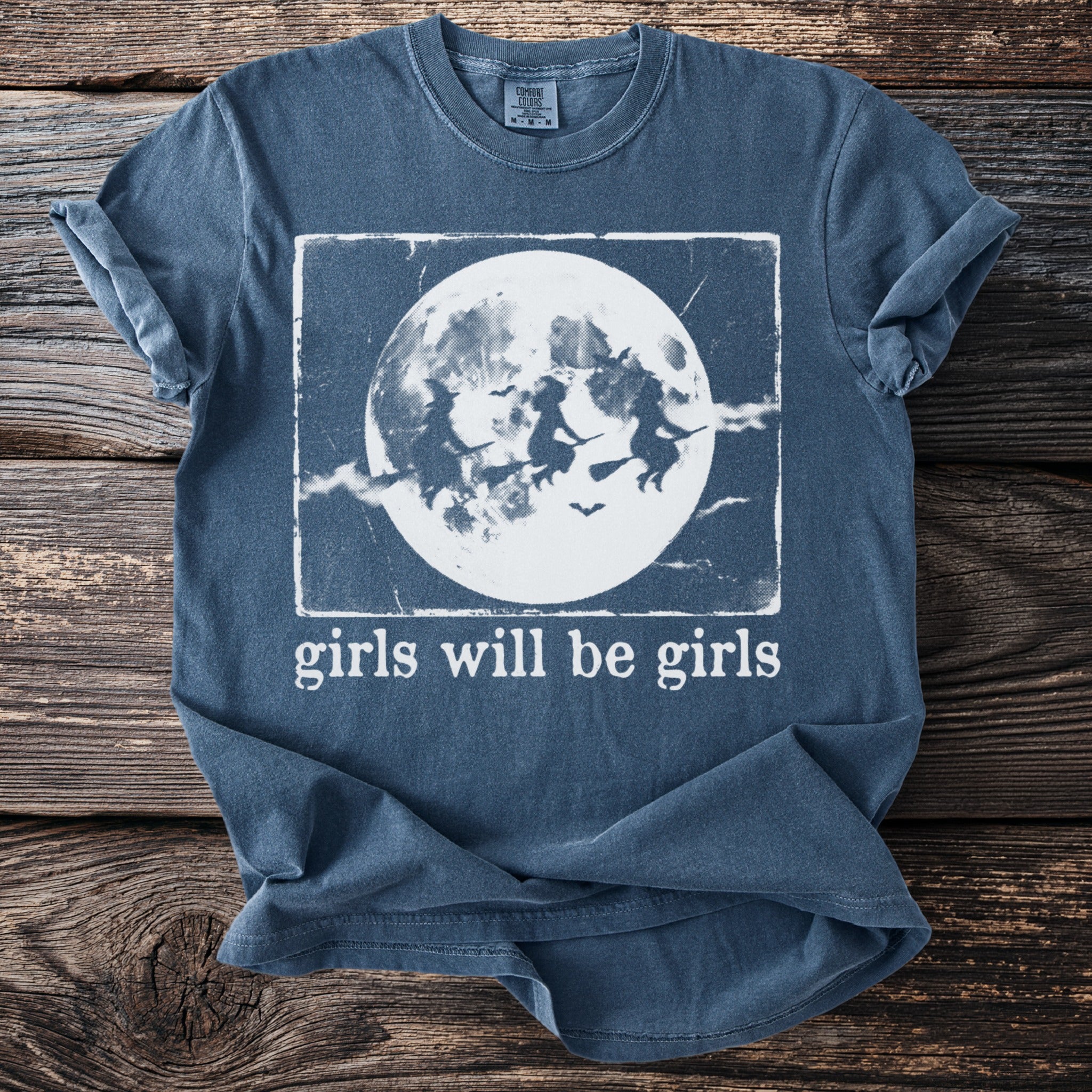 Blue t-shirt with graphic of three witches flying across a moon, text reads "girls will be girls".
