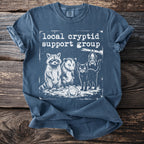 Blue t-shirt with white graphic of raccoon, possum, cat, frog, and bat and text "local cryptid support group".