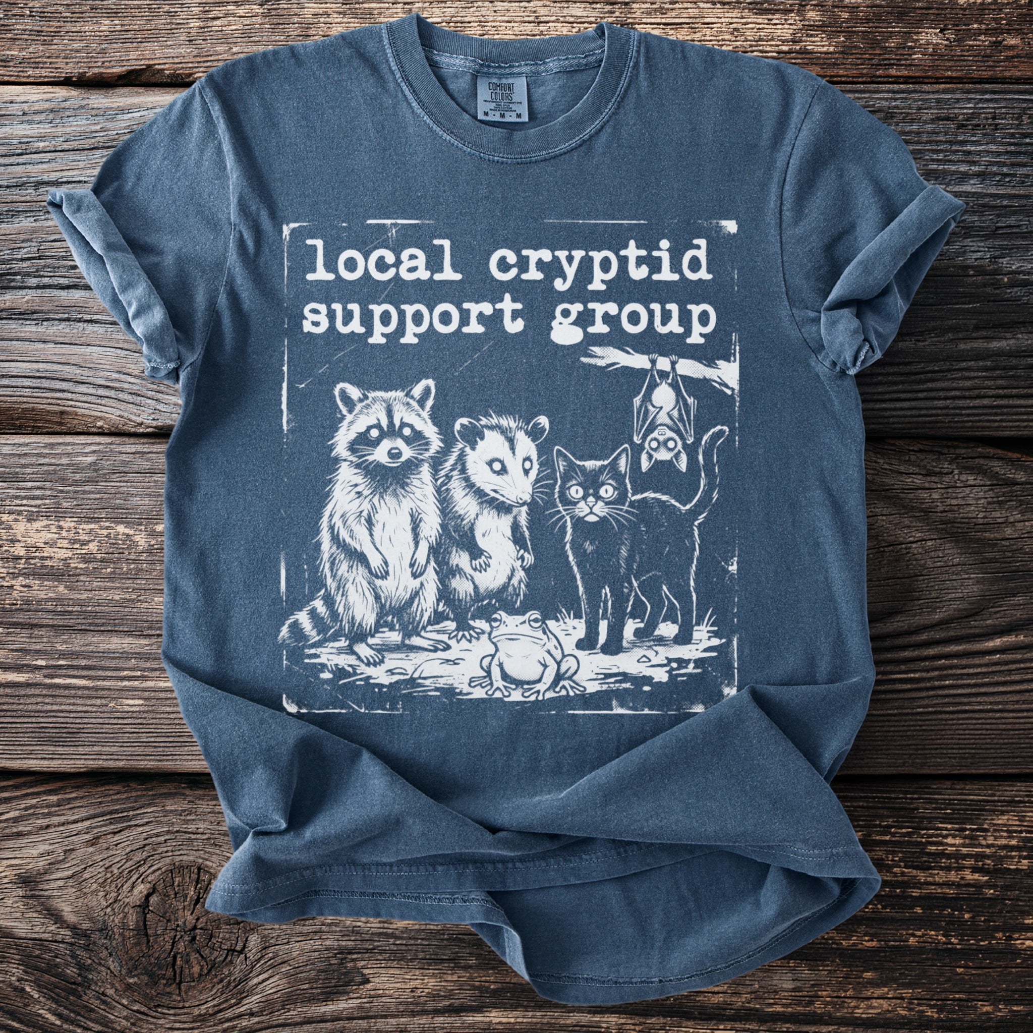 Blue t-shirt with white graphic of raccoon, possum, cat, frog, and bat and text "local cryptid support group".