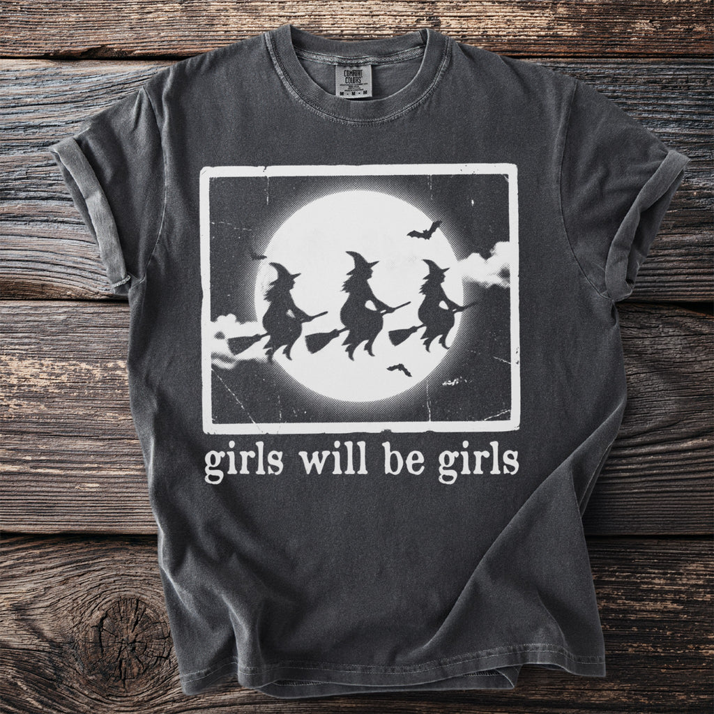 Dark t-shirt with white graphic of three witches flying under a moon and bats, with text "girls will be girls".