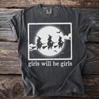Dark t-shirt with white graphic of three witches flying under a moon and bats, with text "girls will be girls".