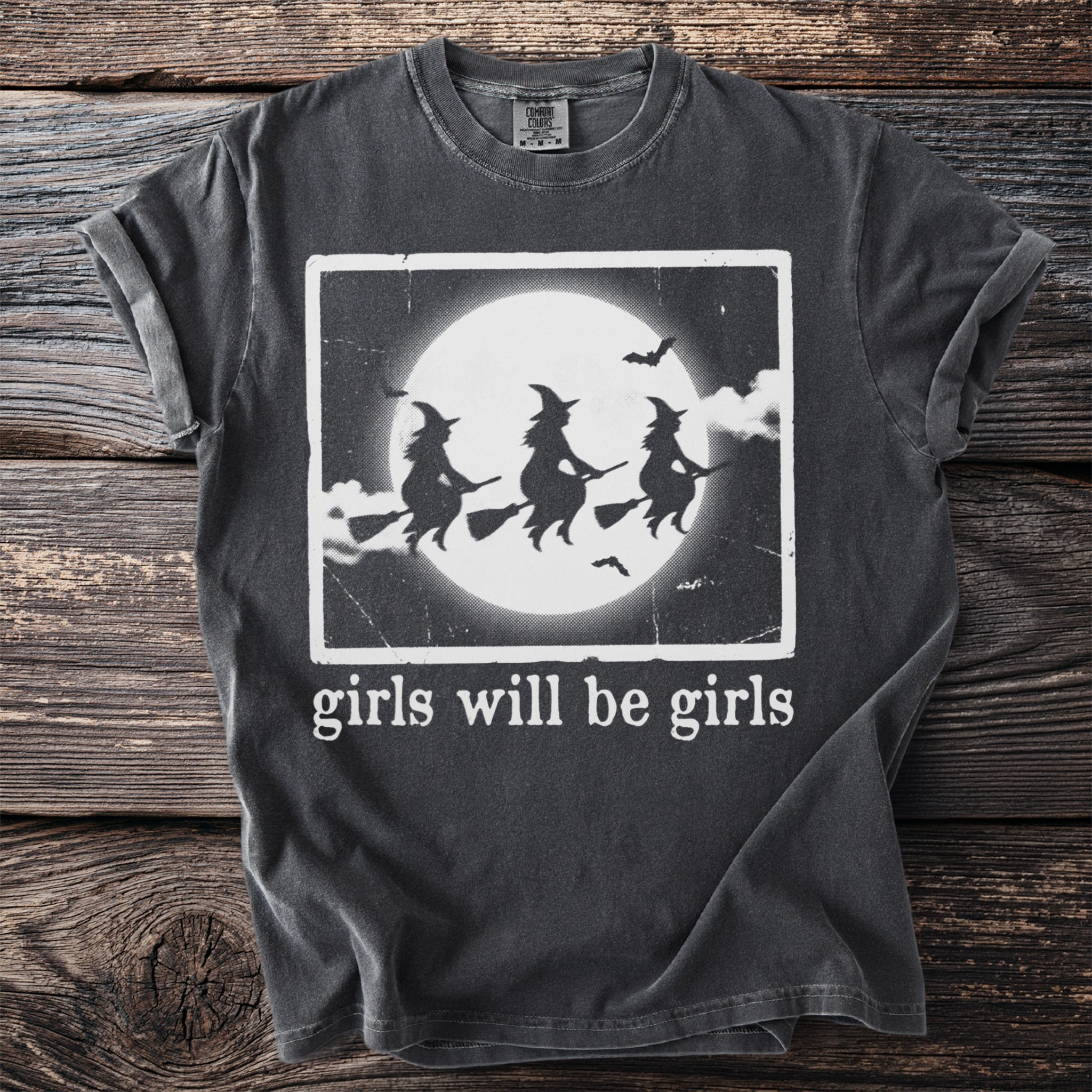 Dark t-shirt with white graphic of three witches flying under a moon and bats, with text "girls will be girls".