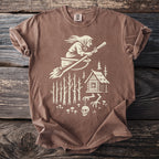 Brown t-shirt with a cream graphic of a witch flying on a broomstick, cabin in woods.