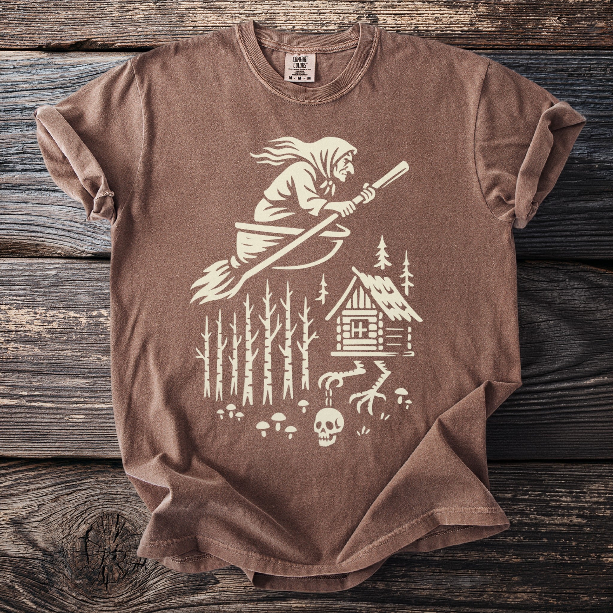 Brown t-shirt with a cream graphic of a witch flying on a broomstick, cabin in woods.