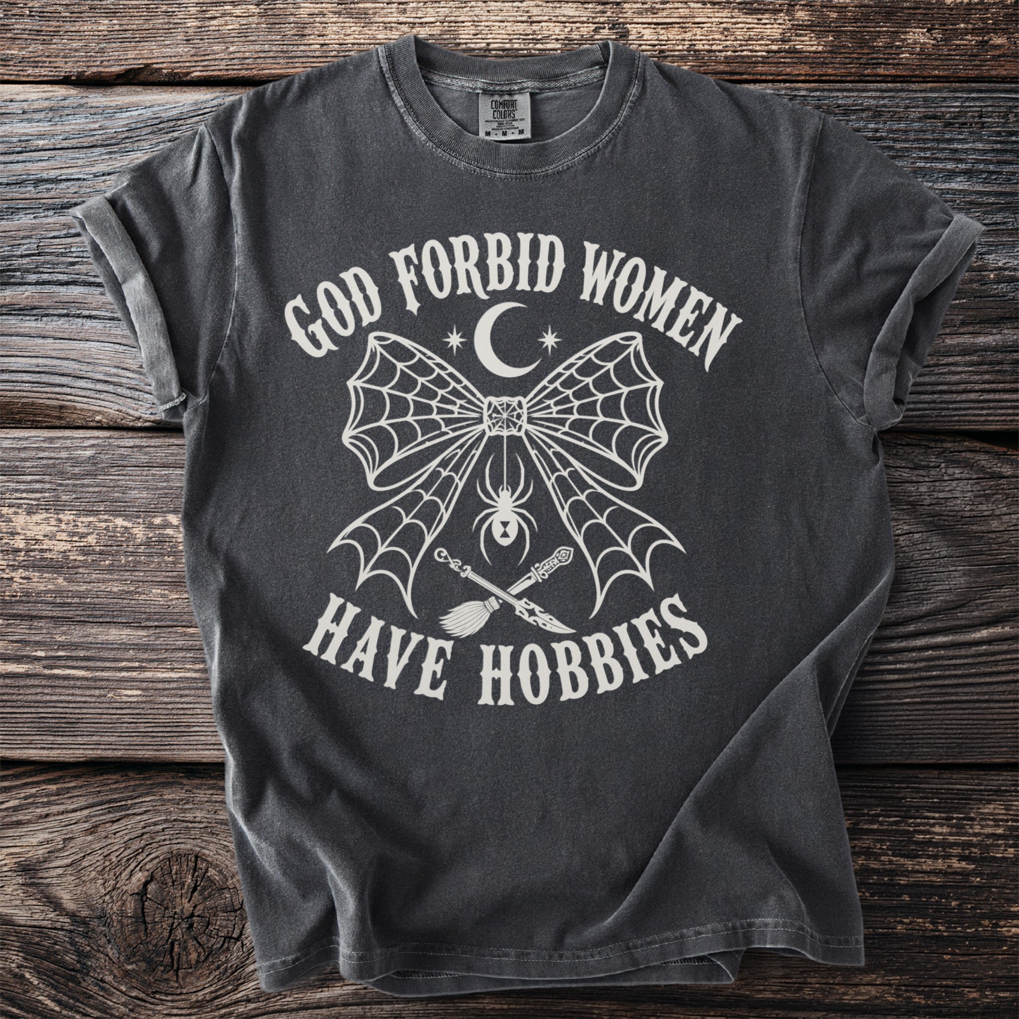 Dark gray t-shirt with white text "God forbid women have hobbies" and gothic graphic.