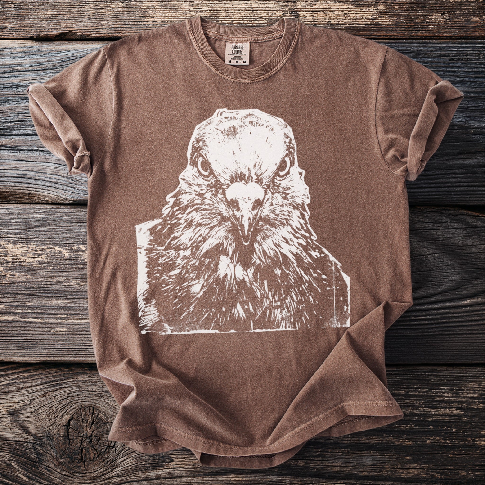 Brown Comfort Colors t-shirt with white graphic of a pigeon's face, size tag visible.