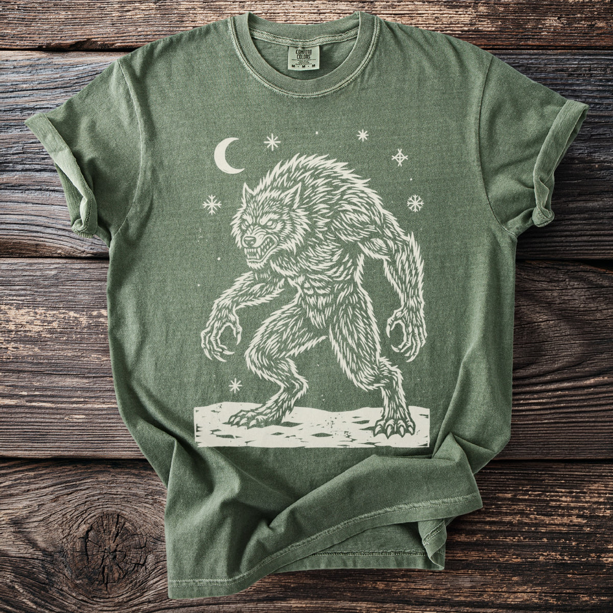 Moss green t-shirt with a cream folk-cut print: a hunched werewolf, teeth bared and claws out, stands in the snow under a crescent moon.
