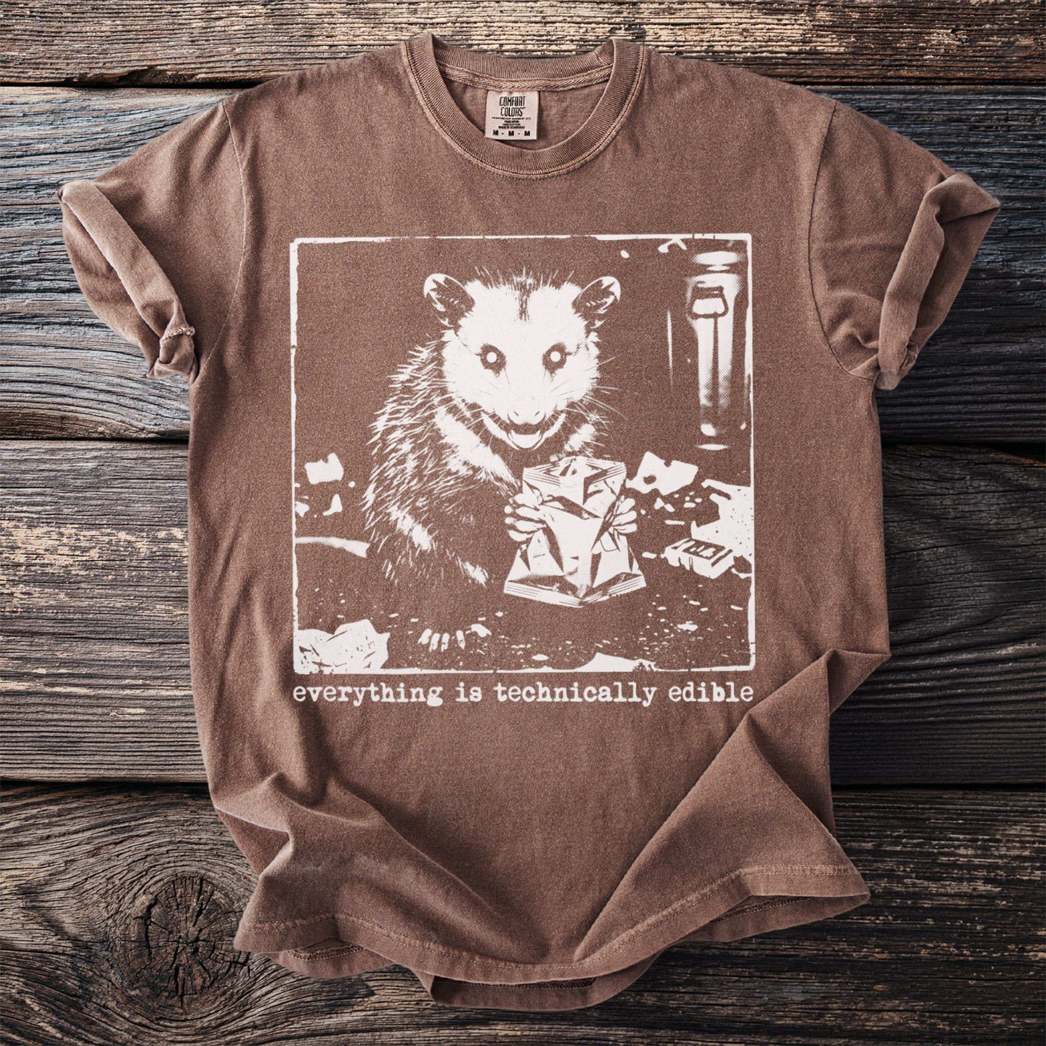 Brown Comfort Colors t-shirt featuring a white graphic of an opossum holding a crumpled bag.