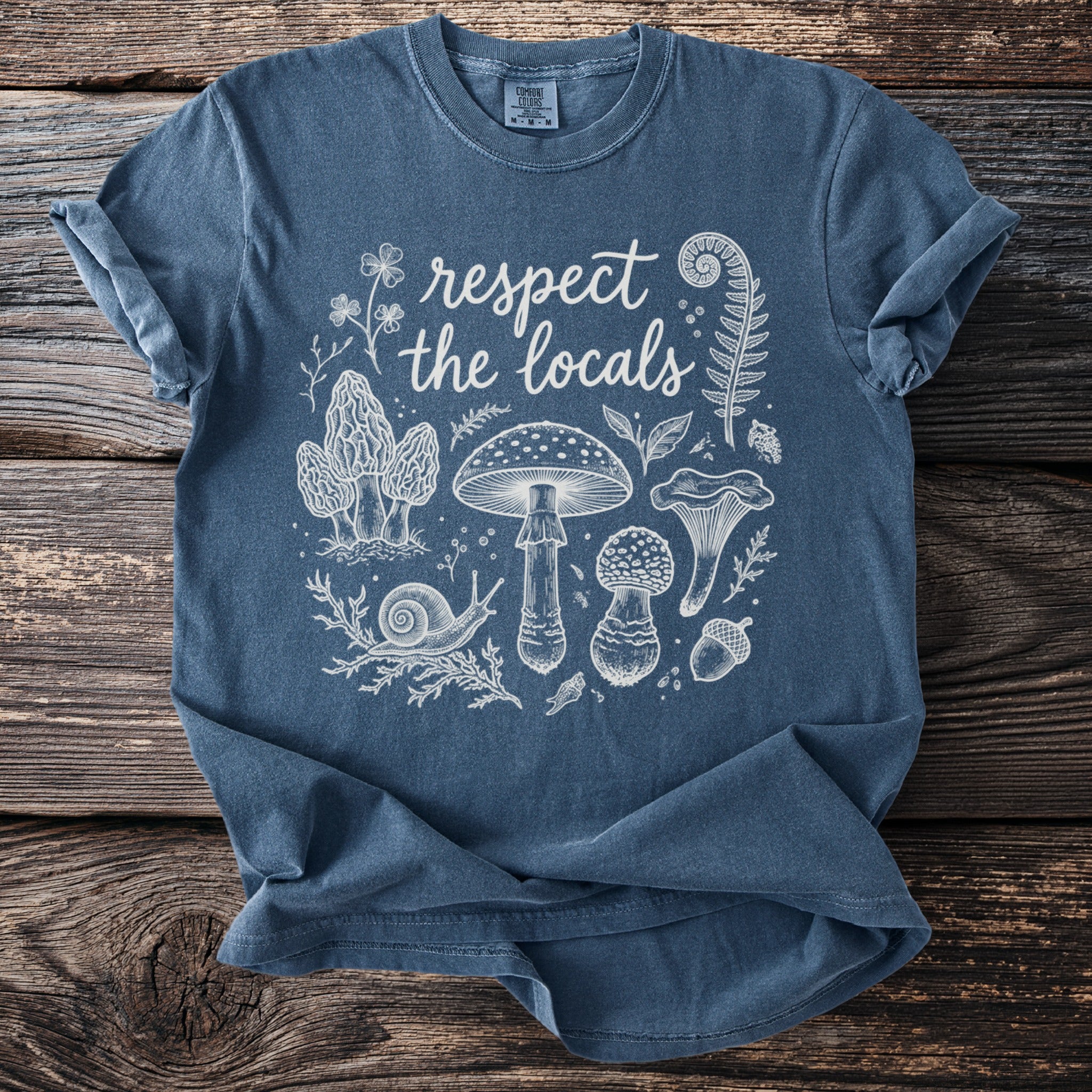 Blue t-shirt with cream graphic of mushrooms, snail, fern, and text "respect the locals".