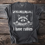 Dark gray t-shirt with a white graphic of a raccoon peeking from a sewer grate and the text "i have rabies".