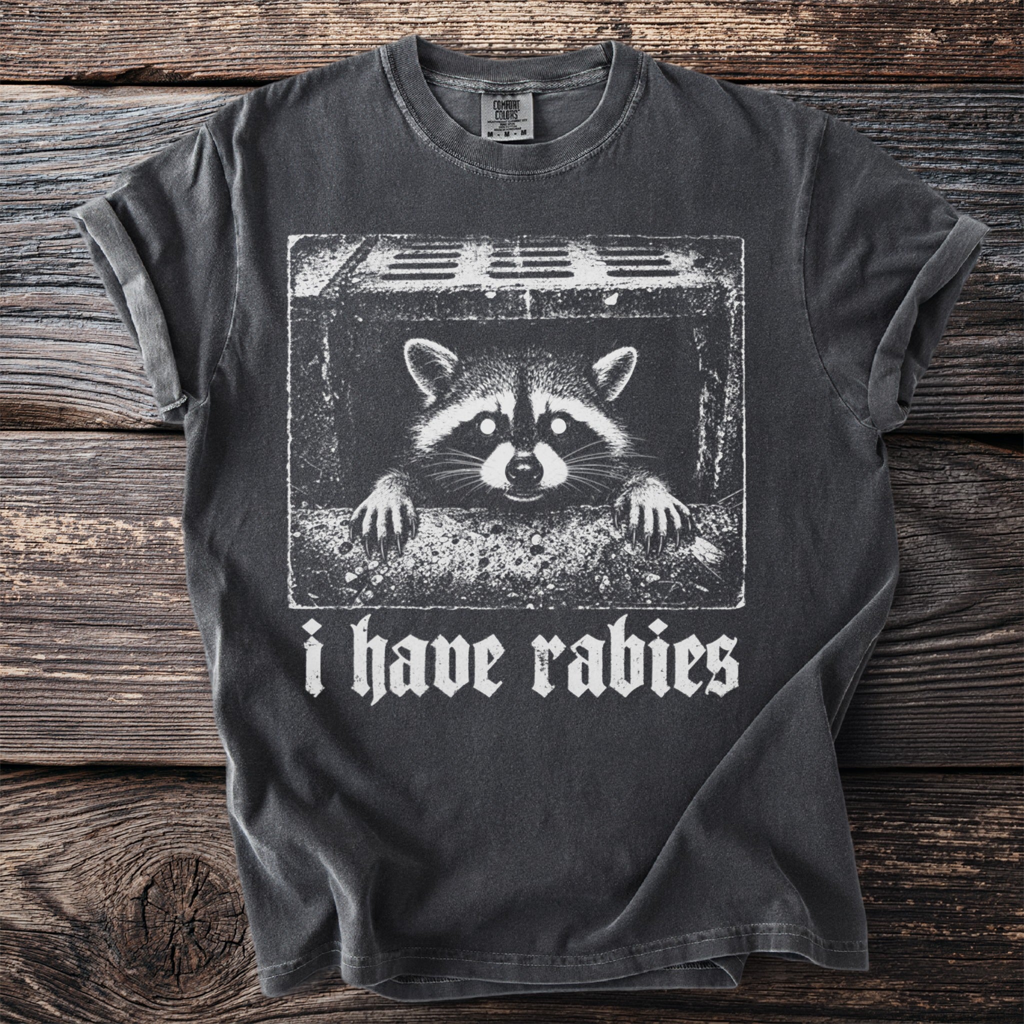 Dark gray t-shirt with a white graphic of a raccoon peeking from a sewer grate and the text "i have rabies".