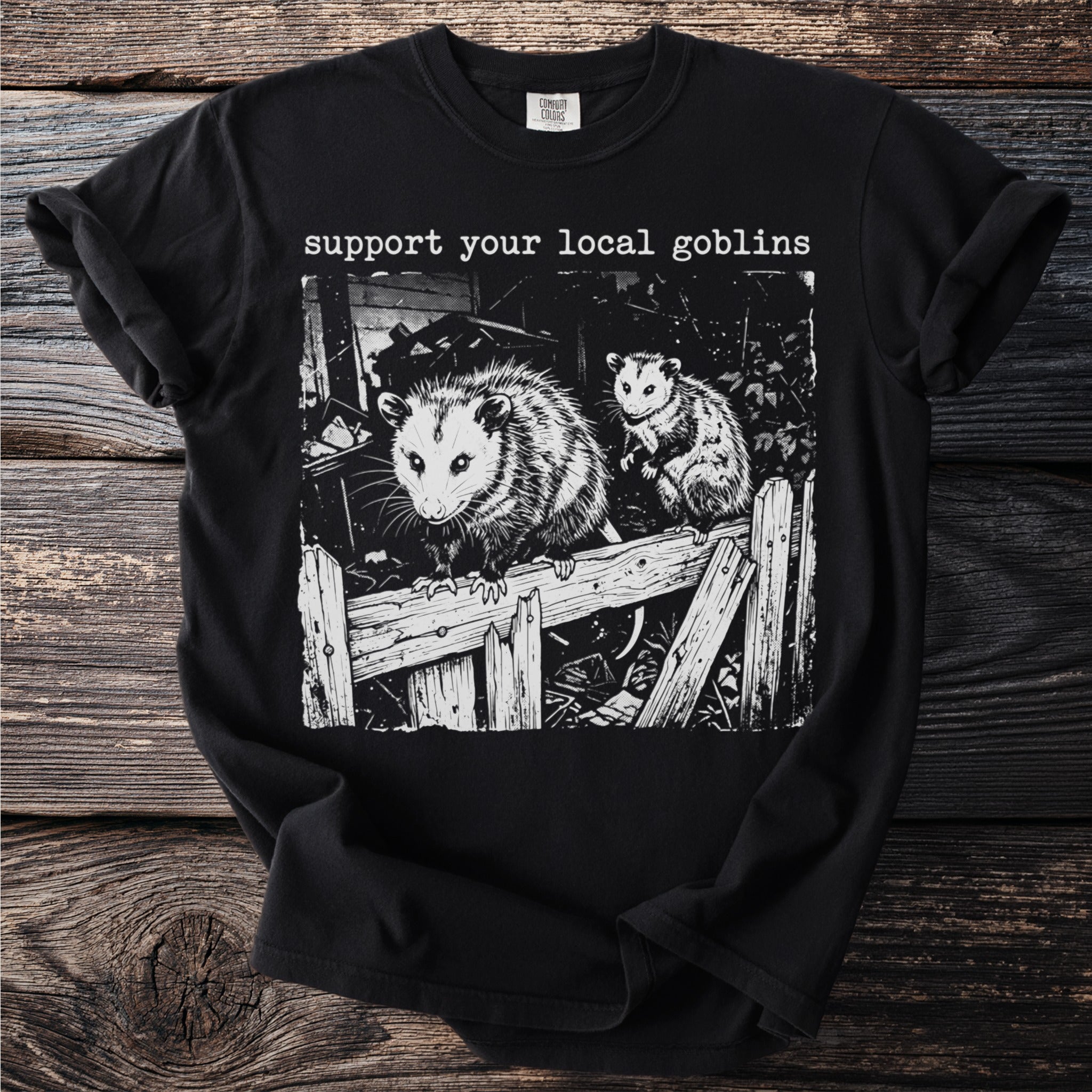 Black t-shirt with "support your local goblins" and opossums on a wooden fence.