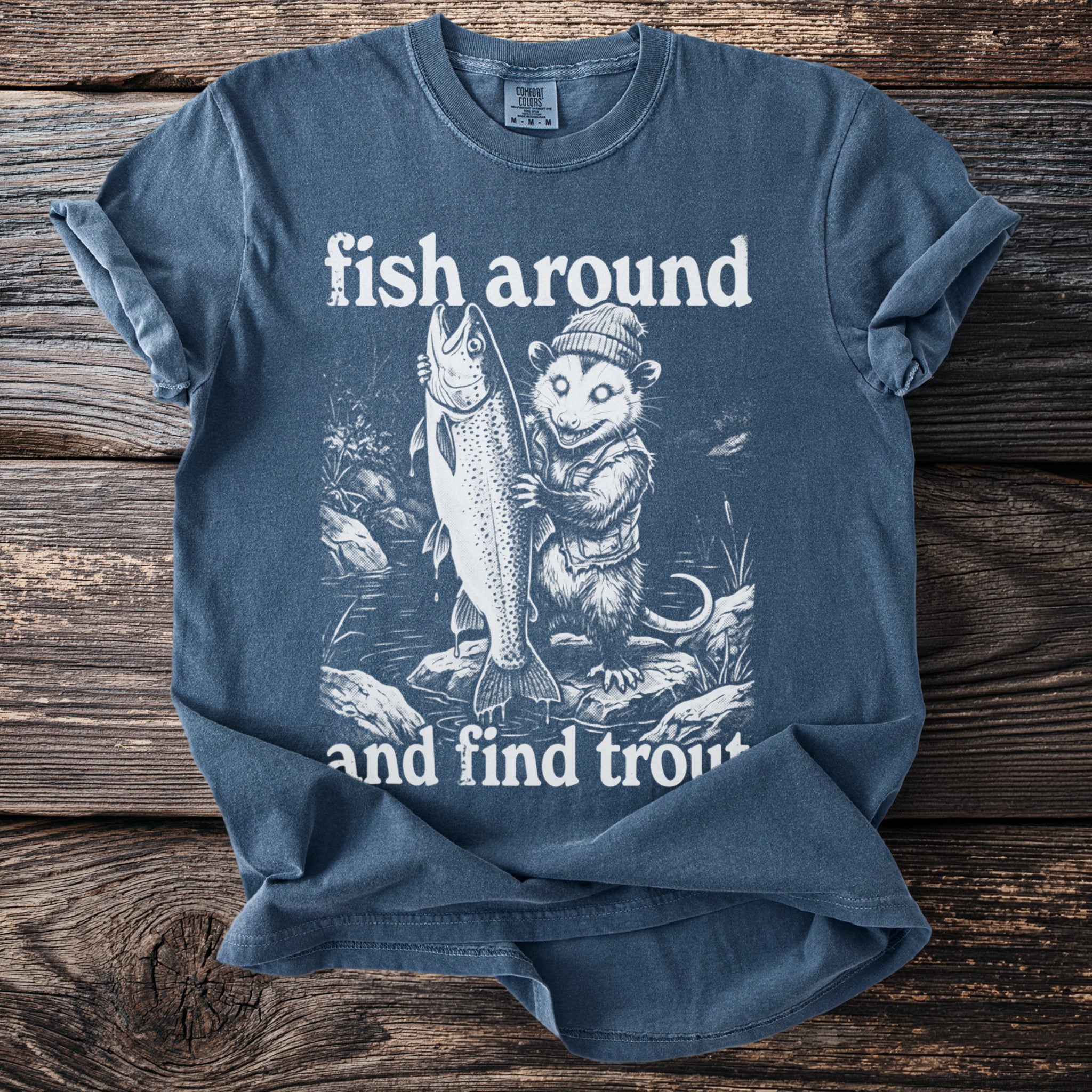 Blue t-shirt graphic with opossum holding large trout, text "fish around and find trout".