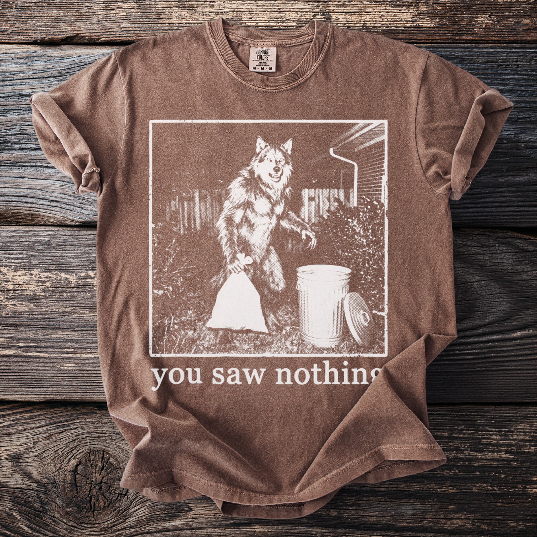 Brown t-shirt with a graphic of a werewolf holding a trash bag next to a trash can and text "you saw nothing".