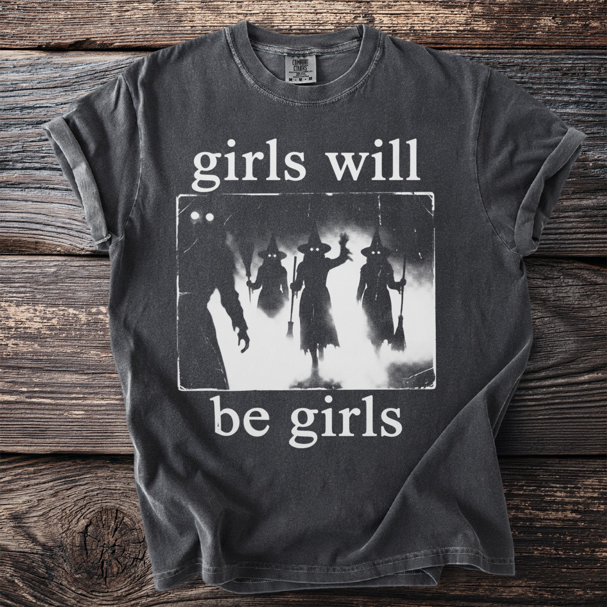 Dark t-shirt with white graphic of silhouettes of three witches and text "girls will be girls."