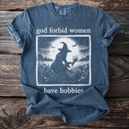 Blue t-shirt with white text and graphic of a witch in a field at night.