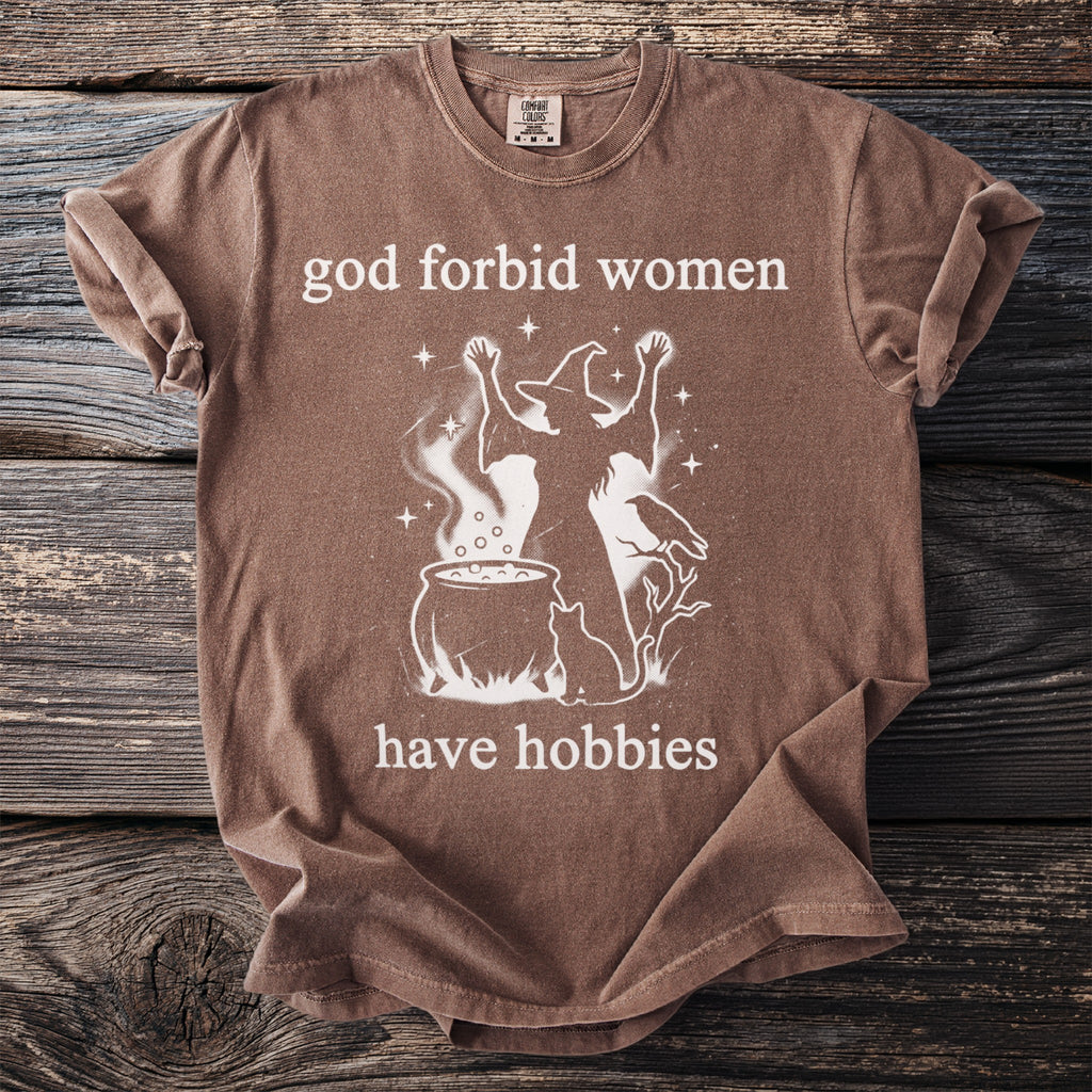 T-shirt with graphic text "god forbid women have hobbies" and witch illustration.