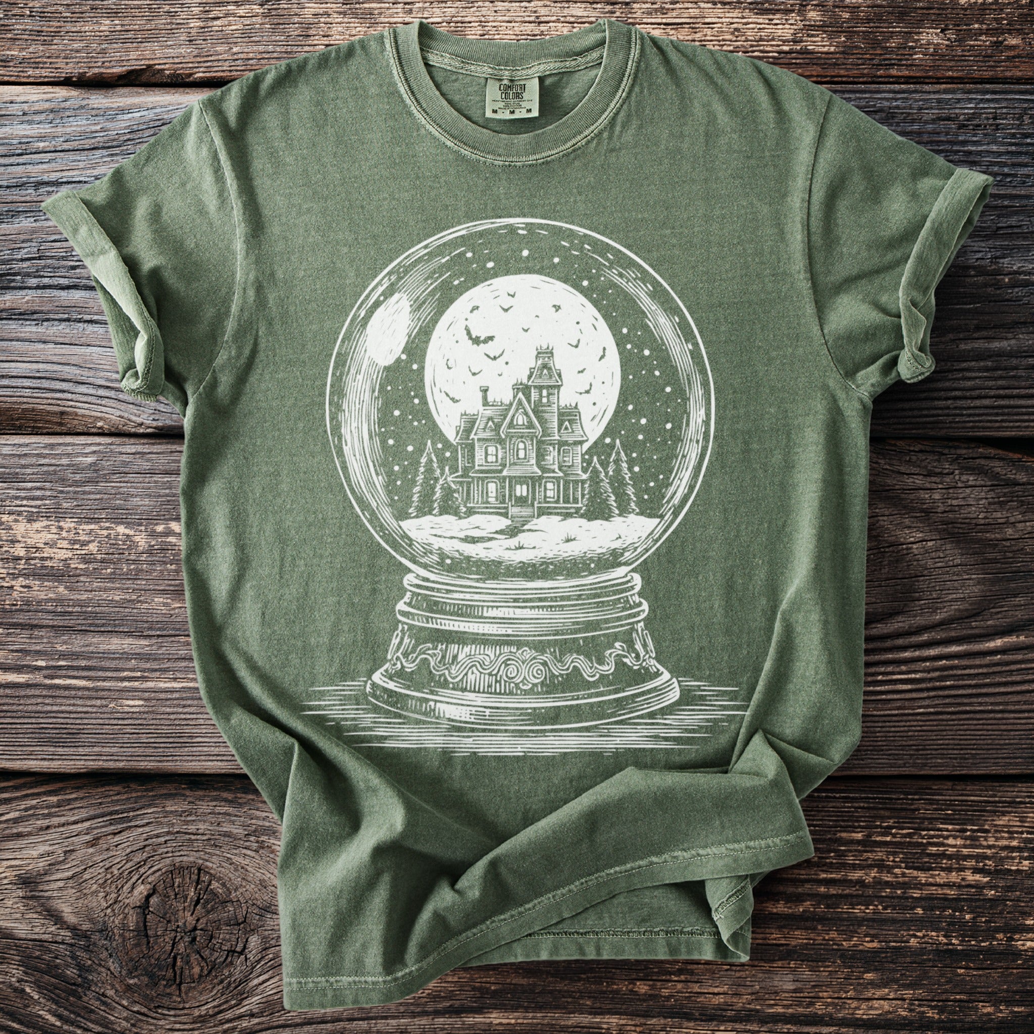 Green t-shirt with a white illustration of a haunted house inside a snow globe.