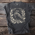 Dark gray t-shirt with a cream linocut style crow surrounded by oak leaves and acorns.