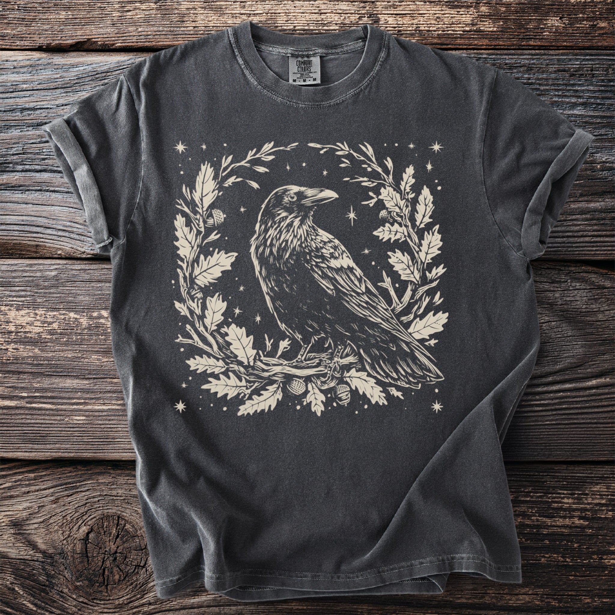 Dark gray t-shirt with a cream linocut style crow surrounded by oak leaves and acorns.